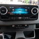 a close-up shot of the dashboard of a 2023 mercedes benz sprinter awd van, focusing on the rectangular landscape-oriented touchscreen in the middle of the dash. the screen is showing audio info, and is flanked on either side by circular air-conditioning vents